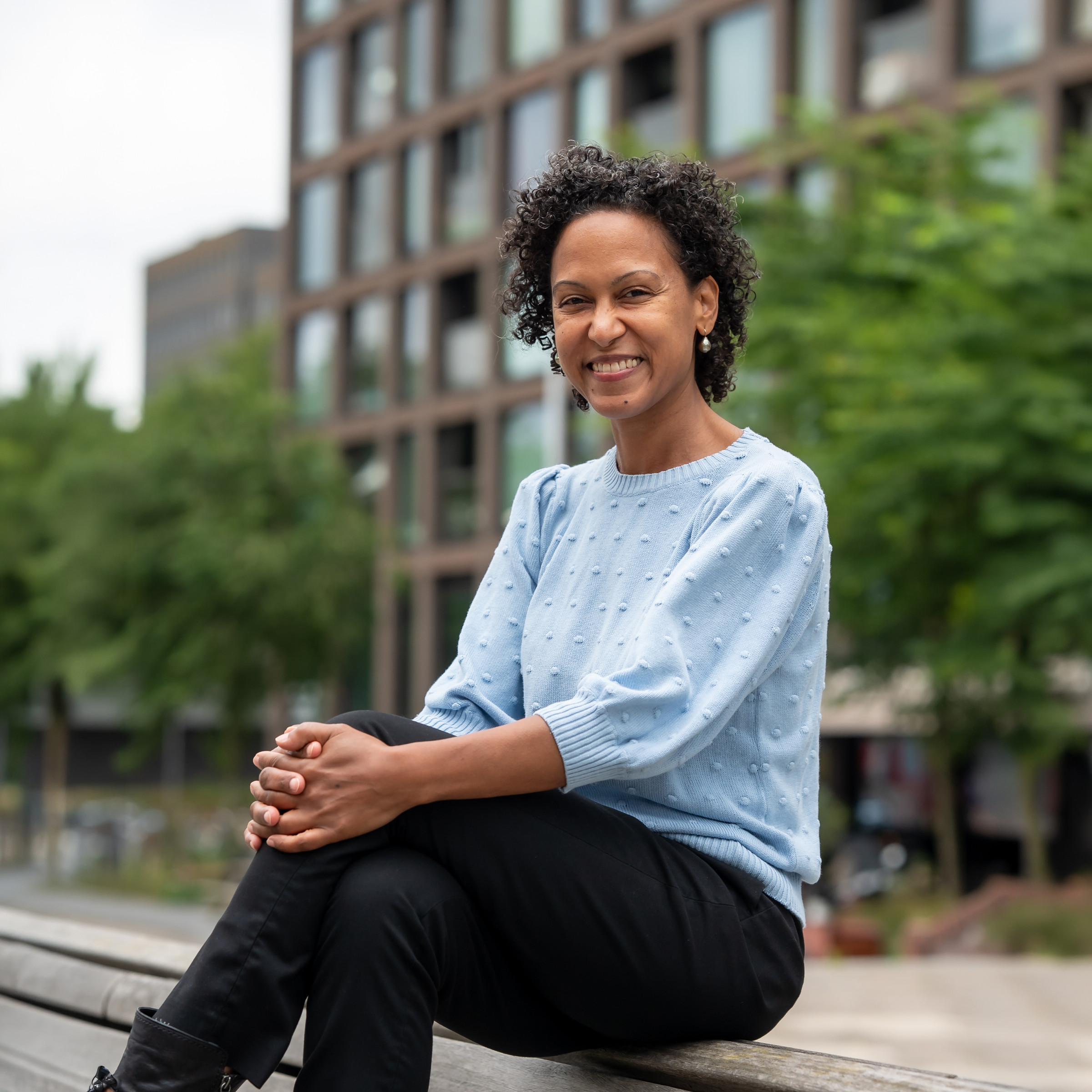 Charissa Maduro zit lachend op een houten bank voor een appartementengebouw.